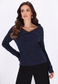 Marineblau gestrickter Pullover mit großem, umgeschlagenem Kragen, gerippter Textur und kunstvollen Zopfmuster, kombiniert mit schwarzen Hosen.
