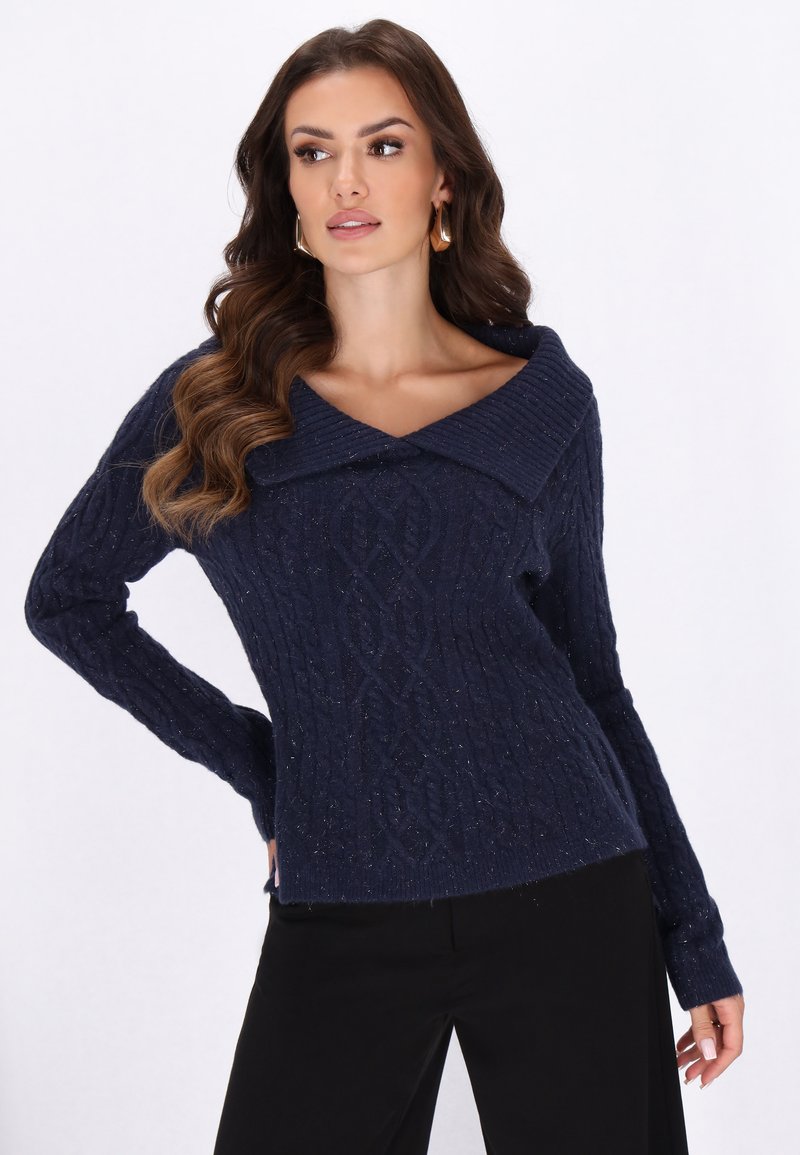 Marineblau gestrickter Pullover mit großem, umgeschlagenem Kragen, gerippter Textur und kunstvollen Zopfmuster, kombiniert mit schwarzen Hosen.