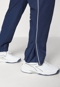Pantaloni da track navy con strisce bianche ai lati, polsini elastici e orli con zip, abbinati a scarpe da ginnastica bianche con accenti blu scuro.