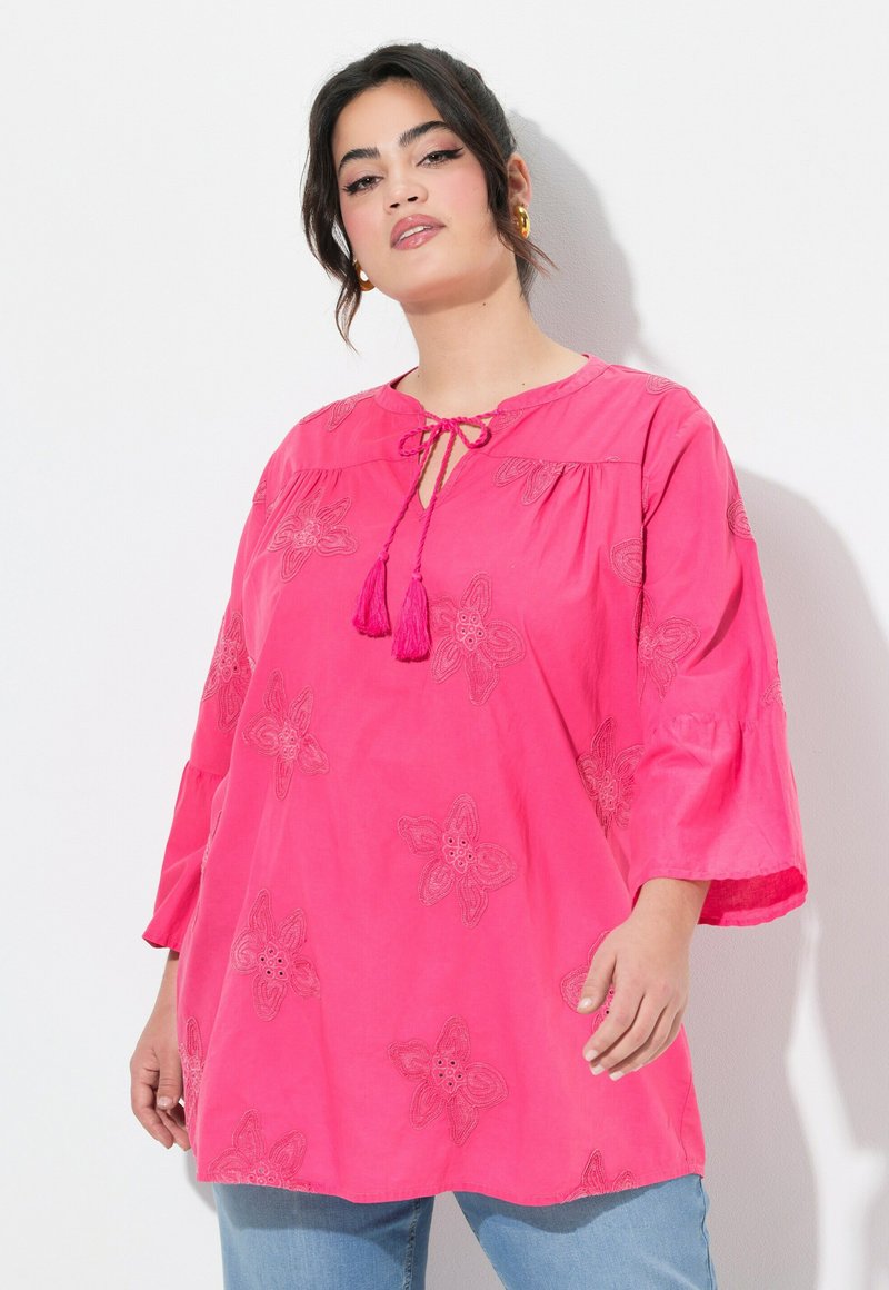 Blusa di cotone rosa con ricami floreali, chiusura con nodo a cravatta e dettagli con nappine. Presenta una vestibilità ampia e maniche a tre quarti a campana.