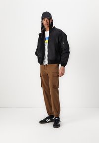 Svart bomberjakke, hvit grafisk t-skjorte med blå og gul tekst, brune cargo-bukser og svarte sneakers med hvite striper.