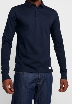Mann trägt ein langärmeliges, dunkelblaues Poloshirt kombiniert mit blauen Jeans und steht vor einem schlichten hellen Hintergrund.