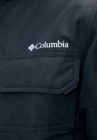 Melna Columbia jaka ar izteiktu krūšu kabatu, izšūti detalizējumi un balts logo kreisajā pusē. Gluda tekstūra.