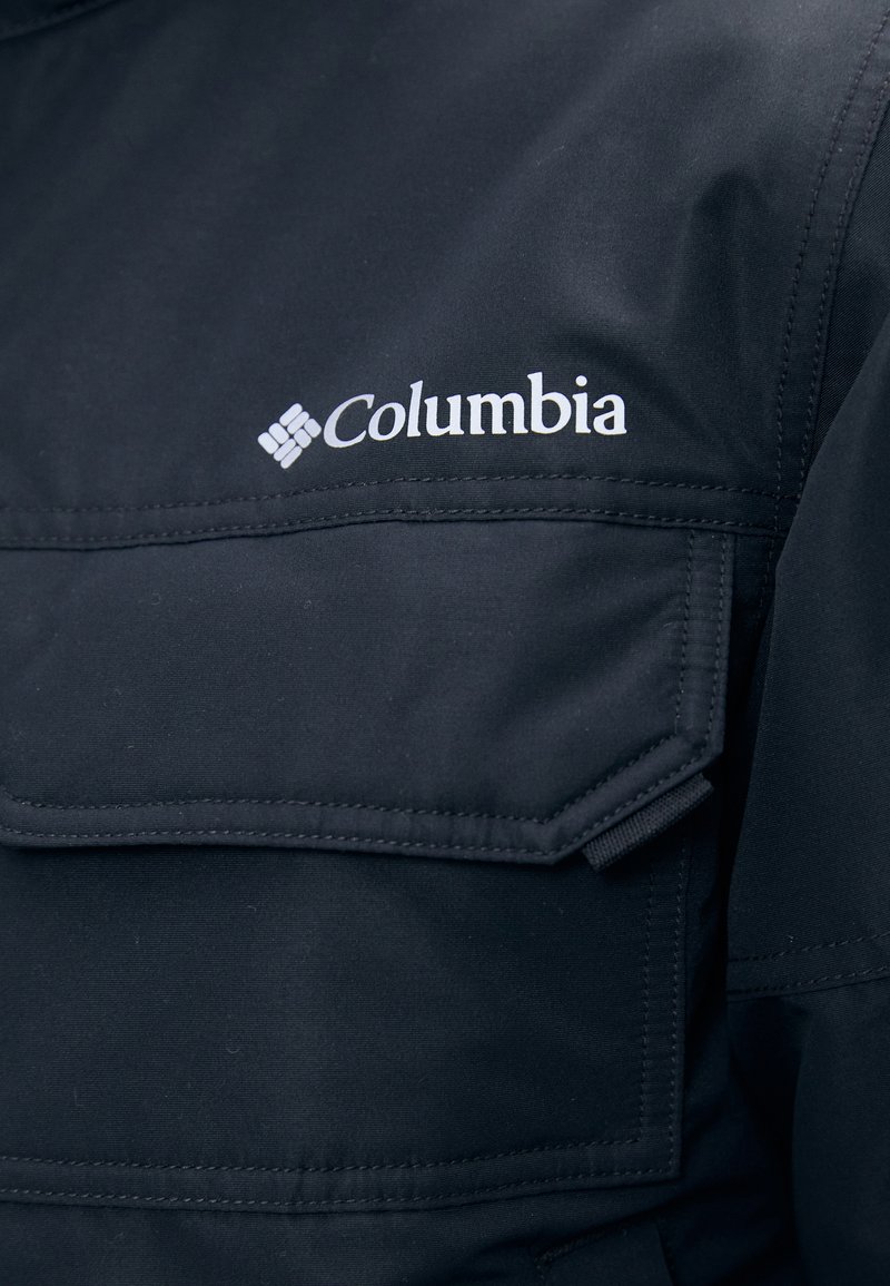 Zwarte Columbia-jack met een opvallende borstzak, gestikte details en een wit logo aan de linkerzijde. Soepele textuur.