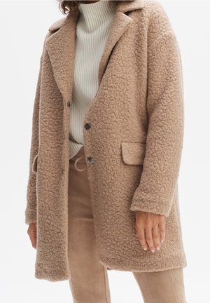 Kvinde iført en beige, struktureret uldfrakke over en hvid, ribstrikket rullekravetrøje og beige bukser med snøre i taljen.