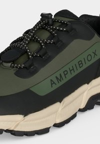Gros plan d'une basket verte et noire avec une semelle épaisse crème, des lacets élastiques noirs et blancs, et le mot "AMPHIBIOX" sur le panneau latéral.