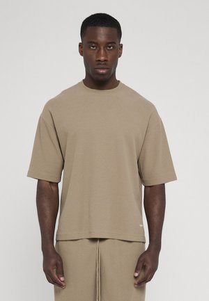 Mand iført beige kortærmet waffle-strikket skjorte og matchende bukser, stående mod en ensfarvet lys baggrund.