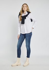 Biała koszula w małe granatowe kropki, połączona z niebieskimi obcisłymi dżinsami i beżowymi botkami za kostkę. Granatowy sweter jest zarzucony na ramionach.