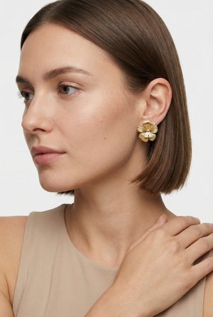 Donna con capelli corti e castani che indossa un top beige senza maniche e un grande orecchino dorato a forma di fiore, toccandosi la spalla con la mano.