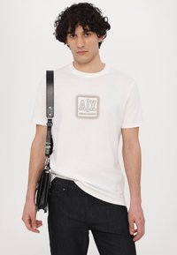 White cotton t-shirt featuring a square graphic with "A|X" and "ARMANI EXCHANGE" in beige. Paired with dark denim jeans and a black bag.