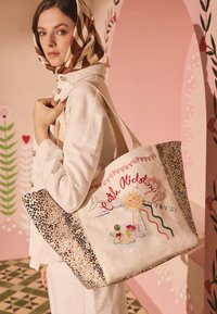 Sieviete gaiši brūnā žaketē un ar rakstainu galvassegu nēsā lielu pirkumu somu ar uzrakstu „Cath Kidston”, saules un varavīksnes dizainu un leopardādas raksta malām.