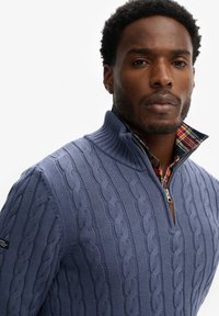 Granatowy sweter w splot warkoczowy z wysokim kołnierzem, z suwakiem. Poniżej częściowo widoczna jest koszula w kratę, co dodaje efektu warstwowości.