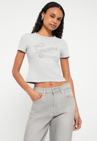 Gray cropped t-shirt with short sleeves, featuring a stylized "Guess" logo in light brown. Paired with light gray jeans and a casual pose.