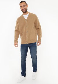 Hellbrauner Strickcardigan mit V-Ausschnitt, vier Knöpfen, zwei Vordertaschen und gerippten Bündchen, kombiniert mit dunkelblauen Jeans und weißen Sneakern.