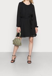 Robe noire à manches longues avec une taille cintrée et un détail de nœud sur le côté. Associée à un sac vert plissé et des sandales à lanières noires.