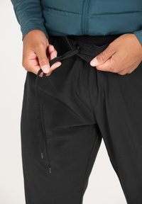 Pantalon de sport noir avec taille élastique et cordon de serrage. Le tissu semble lisse avec une texture subtile, axé sur le confort et l'ajustement.