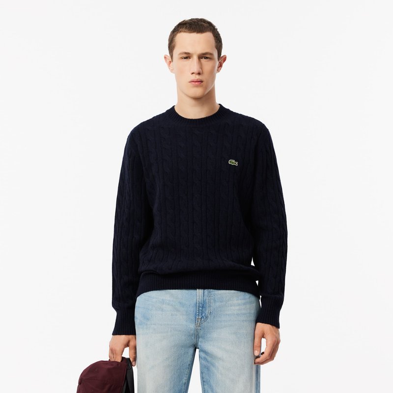 Marineblå kabelstrikket genser med rund hals, ribbestrikkede mansjetter og nederkant. Har en liten grønn logo på brystet. Bært med lyseblå jeans.