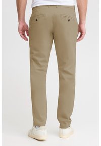 Homme portant un pantalon beige ajusté et des baskets blanches, vu de dos sur un fond clair uni.