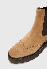 Bottines en suede tan avec des panneaux élastiques sur les côtés, détails brogués au niveau des orteils et semelle en caoutchouc robuste. Texture lisse et lignes épurées.