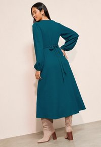 Tamsiai mėlynas ilgomis rankovėmis suknelė su tekstūruotu paviršiumi, aptempta juosmuo, surišta nugaroje, ir plazdanti sijonas, derinama su kremine sukamąja aulinėmis bateliais.