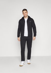 Felpa nera con zip e cappuccio regolabile, abbinata a pantaloni della tuta neri. Una maglietta bianca sotto e scarpe da ginnastica bianche completano l'outfit.