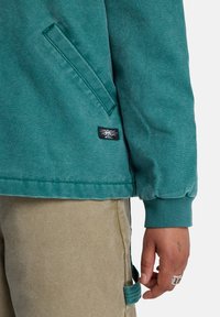 Teal katoenmengsel jack met een zijzak, ribbelmanchetten en een zwart logo-opkleefstuk. Draagt met lichtbeige broek.