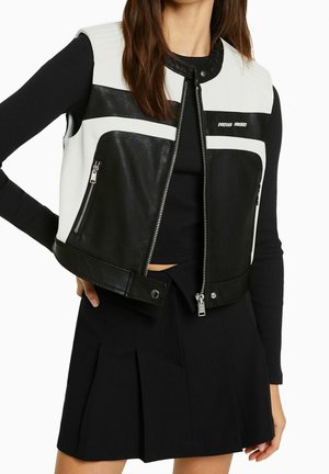 Woman wearing black and white leather sleeveless zip vest over black long-sleeve top and black pleated skirt.