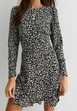 Frau trägt ein schwarzes, langärmliges Kleid mit kleinem weißem Blumenmuster und Rüschensaum, accessoriert mit mehrlagigen silbernen Halsketten.