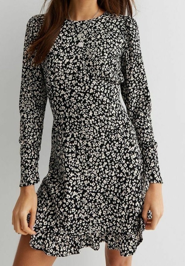 Femme portant une robe noire à manches longues avec un petit imprimé floral blanc et un ourlet volanté, accessoirisée avec des colliers argentés superposés.