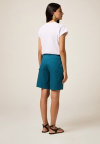 T-shirt blanc à manches courtes associé à un short bermuda teal avec deux poches à l'arrière et des détails de couture subtils. Sandales noires à lanières.
