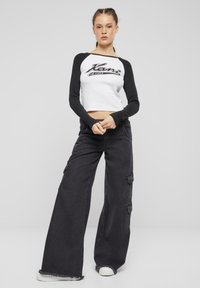 Černý dlouhý rukáv crop top s bílou grafikou na přední straně, spárovaný s volnými černými cargo kalhotami s bočními kapsami a třepenými lemy.