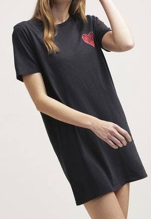 Kobieta ubrana w czarną sukienkę z krótkim rękawem z naszywką w kształcie czerwonego serca na piersi, stojąca na jasnym, jednolitym tle.