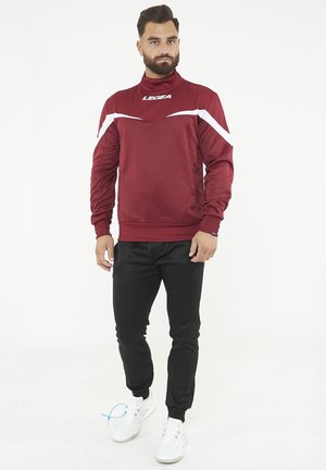 Uomo che indossa una maglia sportiva a maniche lunghe color granata con strisce bianche, pantaloni da jogging neri e scarpe da ginnastica bianche, in piedi contro uno sfondo semplice.