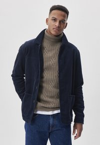 Jachetă din bumbac cord cu guler, de culoare bleumarin, cu închidere prin buton și două buzunare frontale, purtată peste un pulover cu gât înalt de culoare bej, cu model cu nervuri.