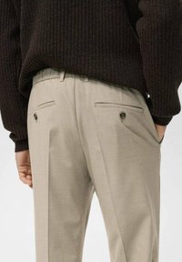 Pantalones beige a medida con pierna recta, doble bolsillo trasero y detalles de costura sutiles, combinados con un suéter oscuro de canalé.