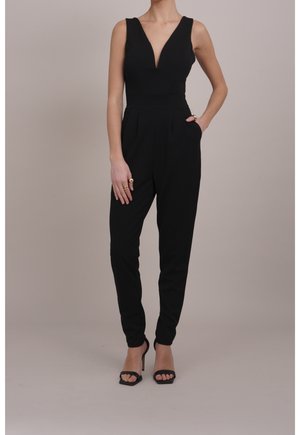 Kvinde iført en ærmeløs sort jumpsuit med dyb V-udskæring og tilspidsede bukser, kombineret med sorte højhælede sandaler og gyldne ringe.