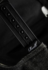 Correa de tela negra con un tejido texturizado, que presenta una hebilla de plástico negro liso y una etiqueta blanca "Reell" cosida en una lona tejida.