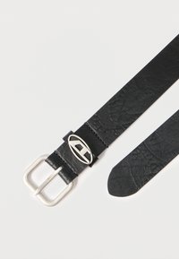 Melns ādas josta ar sudraba taisnstūrveida sprādzi un ovālu akcentu. Tai ir teksturēts raksts un sašaurināts gals.