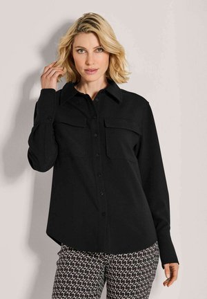 Blonde vrouw in een zwarte overhemd met knopen en borstzakken, met een patroon van zwart-wit broek, terwijl ze haar haar aanraakt.