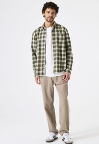 Camisa de cuadros verde y crema, abotonada, superpuesta a una camiseta blanca; combinada con pantalones de pana beige y zapatillas blanco y negro.