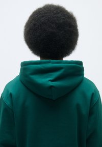 Felpa verde con cappuccio, tessuto morbido, vestibilità comoda, vista posteriore che mostra i dettagli del cappuccio e il girocollo arrotondato, strutturata con un'acconciatura morbida visibile.