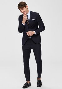 Marinblå kostym med en slim fit-design, enkel knappstängning, ljusblå skjorta, randig slips och en mönstrad näsduk. Svarta loafers.