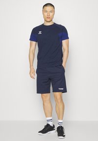 Mężczyzna stoi, ubrany w granatową sportową koszulkę z krótkim rękawem i pasujące spodenki, białe skarpetki z granatowymi paskami oraz czarne tenisówki.