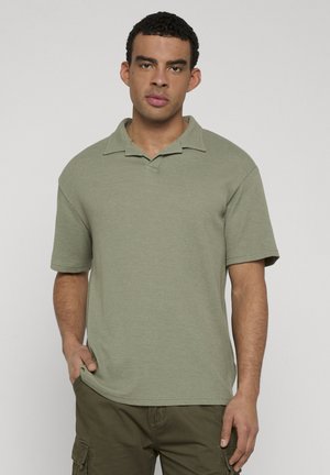 Mann iført lys grønn kortermet poloshirt og olivengrønne cargobukser, stående med en hånd i lommen mot enkel bakgrunn.