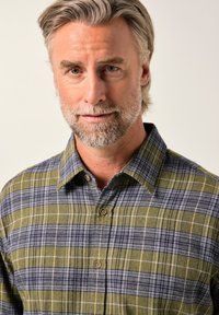 Muž středního věku se šedými vlasy a vousy, oblečený v kostkované košili v zelené a modré barvě, mírně se usmívající na jednorázovém pozadí.