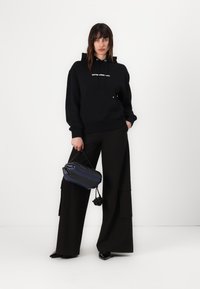 Felpa nera con cappuccio e logo frontale, abbinata a pantaloni neri larghi. Il modello tiene in mano una borsa a righe blu e nere.