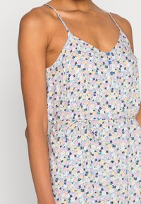 Nainen, joka pukeutuu pastellinsävyiseen kukkakuviolliseen hihattomaan mekkoon, jossa on ohuet olkaimet, napit rinnassa ja sidottava vyötärövyö.