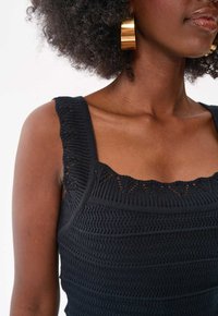 Zwarte gebreide tanktop met een vierkante halslijn met scalloped randen, textuurstof en brede schouderbanden. Gouden hoepeloorbellen zichtbaar.