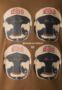 Vier stilisierte Vintage-Boxhelme mit "HUGO" in Rot oben, angeordnet in einem Raster über dem Text "BOXING ACADEMY 93" auf braunem Stoff.