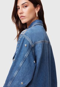 Giacca di jeans con dettagli in borchie argentate sulle spalle, tasche con pattina e lavaggio blu classico. Presenta una vestibilità rilassata e un tessuto robusto.